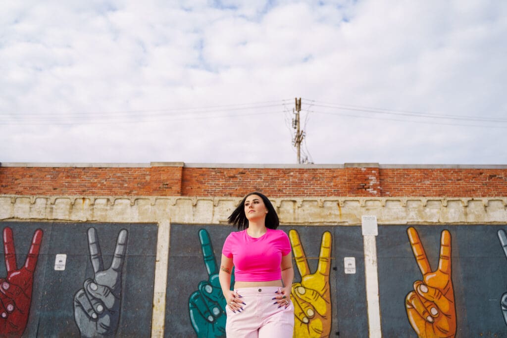 Senior in a bright pink outfit by peace sign graffiti in Franklinton Columbus, Ohio.