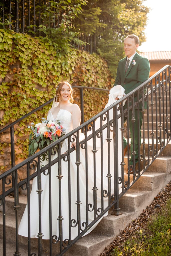 The groom holds the bride's train as she walks down the stairs.