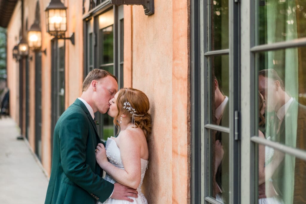 Bride and groom share a kiss against a wall with their reflection in the window.