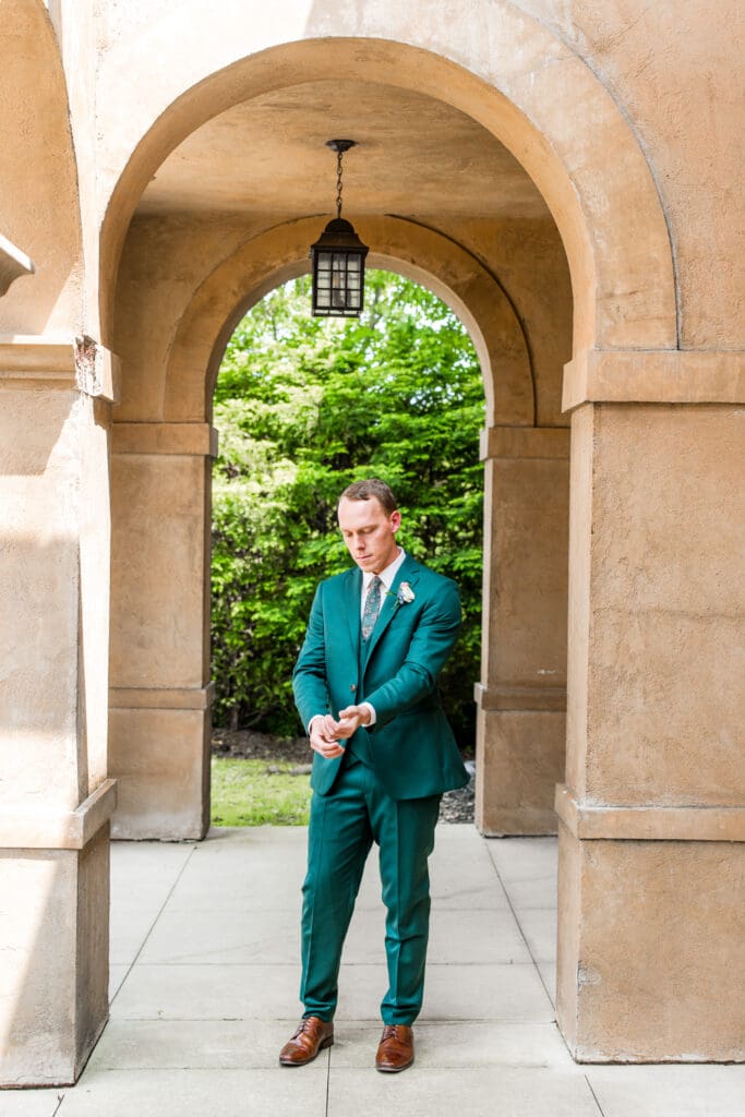 The groom fixes his sleeves under an arched wall.
