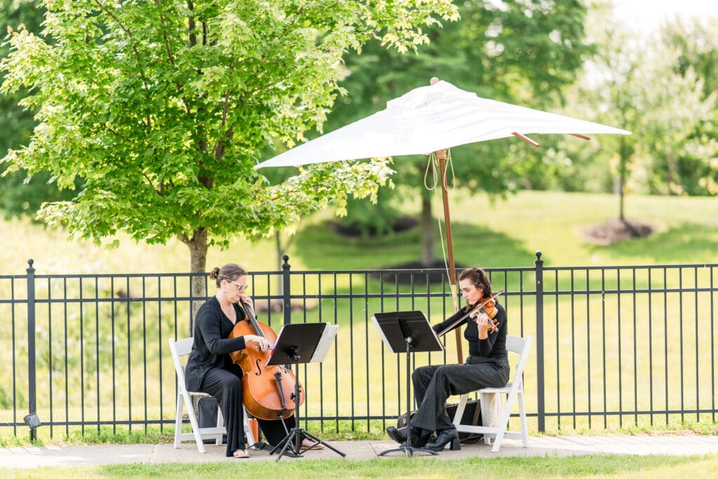 Two people playing string instruments under an umbrella at an outdoor wedding ceremony.