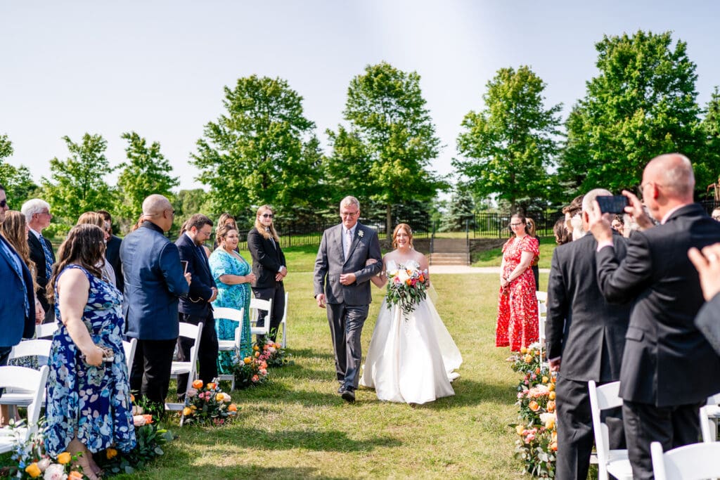 The bride and her father walk down the aisle while her guests watch.
