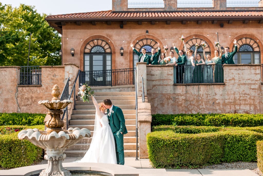The bride and groom kiss in front of a staircase and fountain while their wedding party cheers in the background.