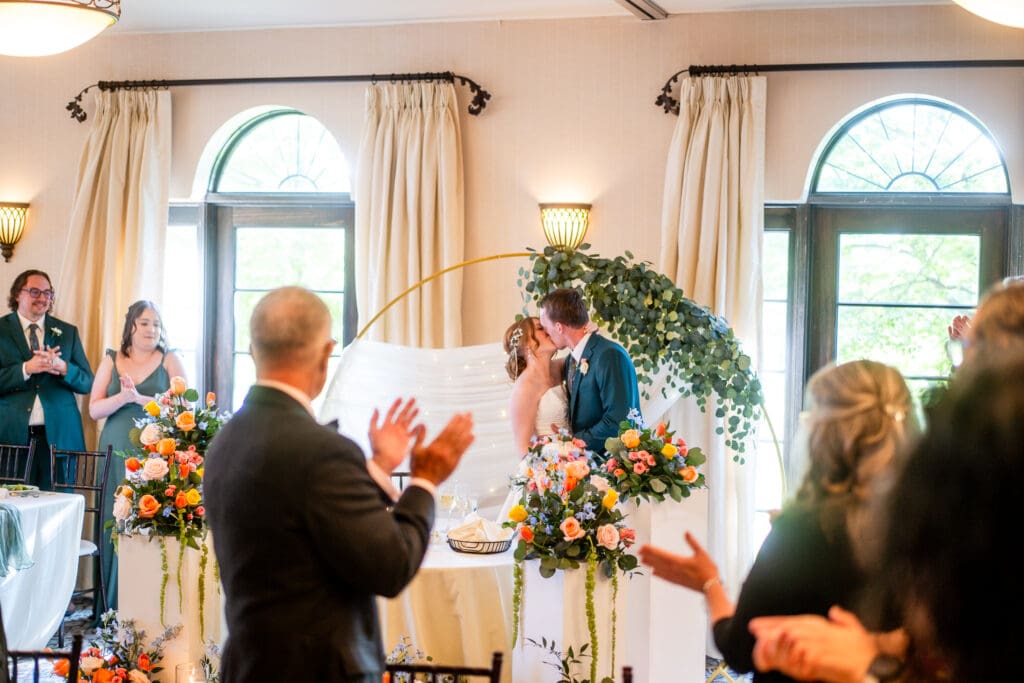 Bride and groom kiss at their sweetheart table while guests cheer.