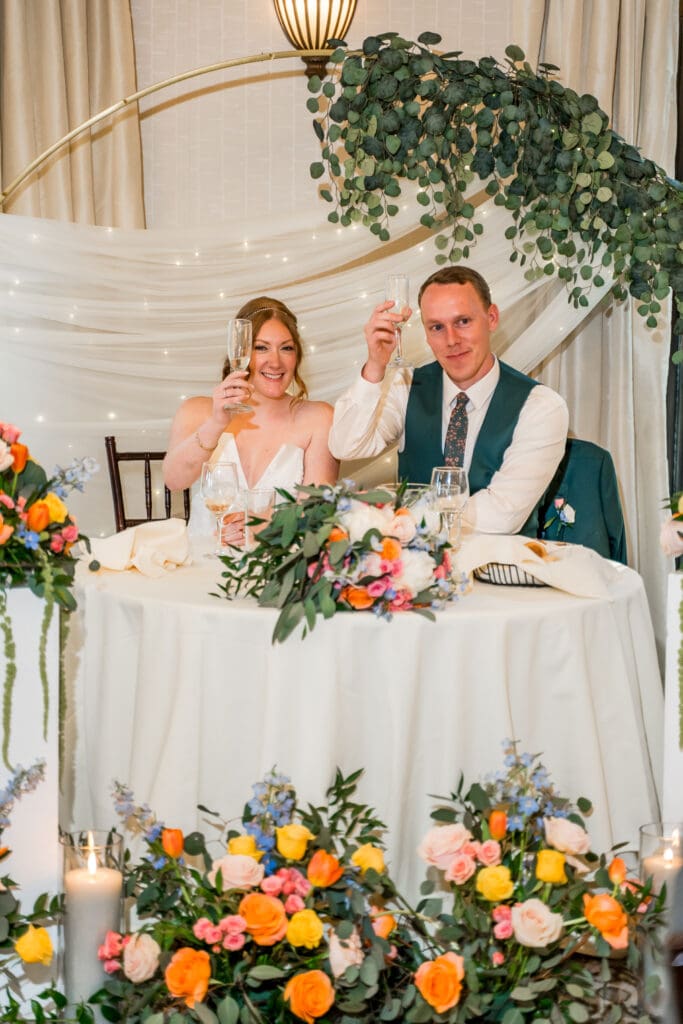 Bride and groom hold out their champagne glasses in a cheers at their colorful sweetheart table.