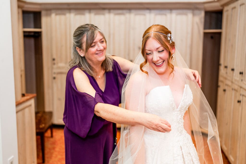 A bride laughs with her mom as her mom puts her veil around her shoulders.