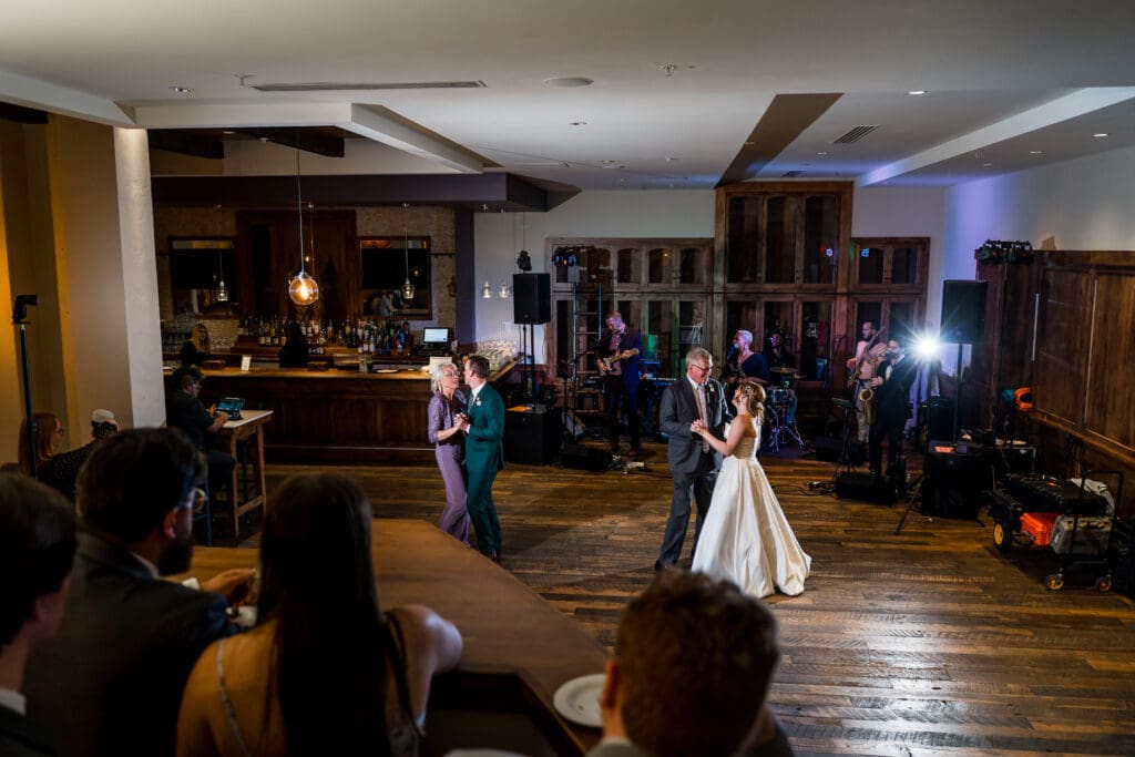 The bride and her father and the groom and his mother dance on the dance floor as the band play and guests watch.