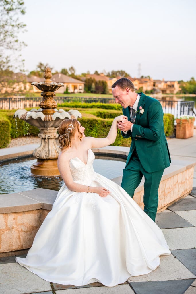 The groom takes the bride's hand as she sits on the edge of a fountain.