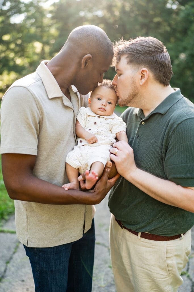 Two dads kiss their baby boy on the head as they hold him.
