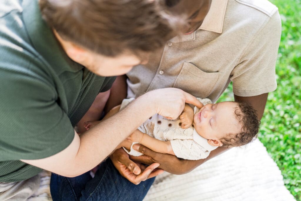 Two dads hold their baby boy in a picnic setting outdoors.