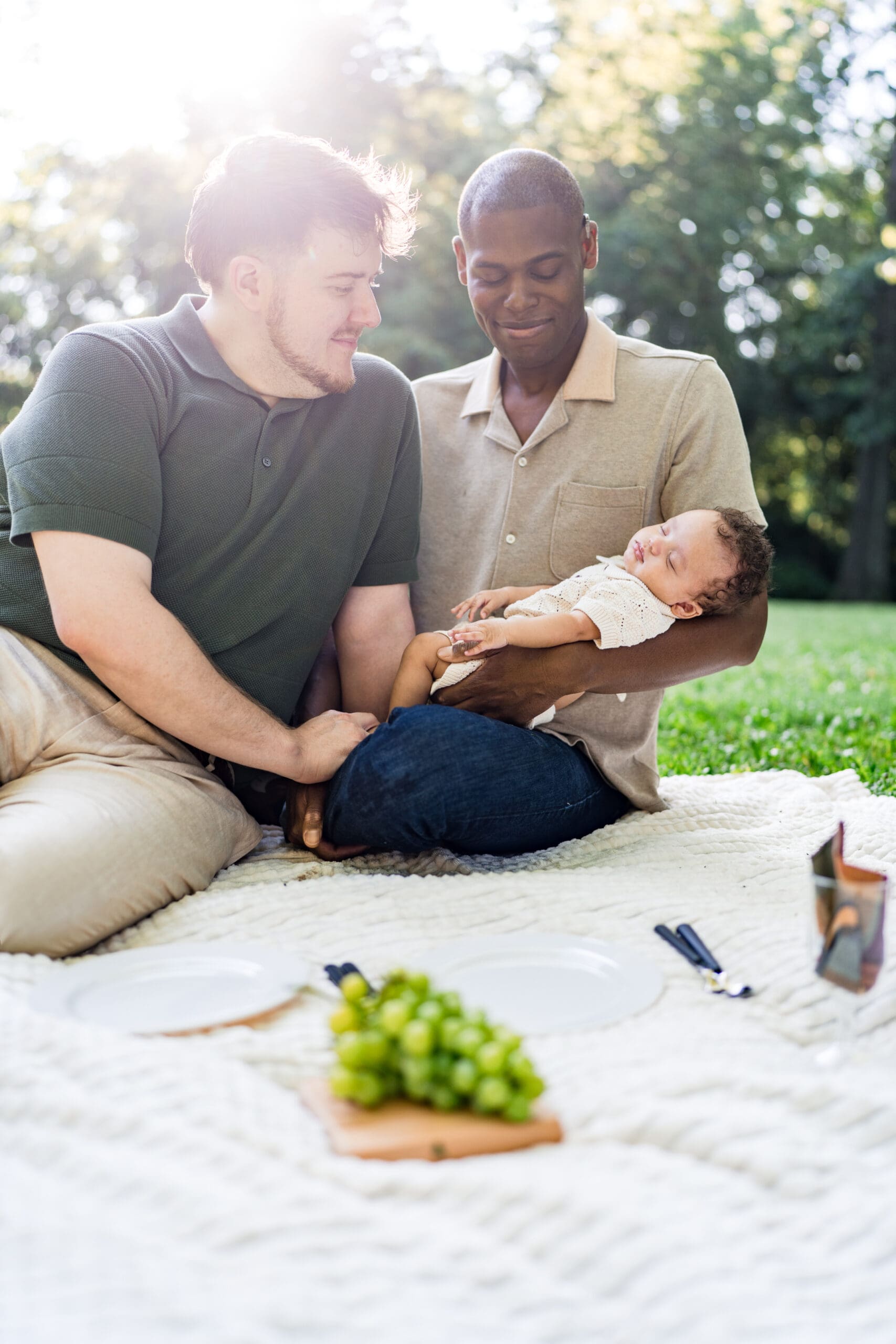 Two dads hold their sleeping baby son in an outdoor picnic setting.