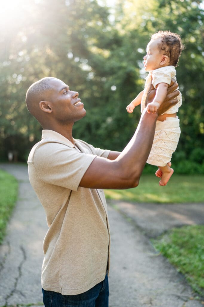 A dad hold his son up and looks at him in a park.