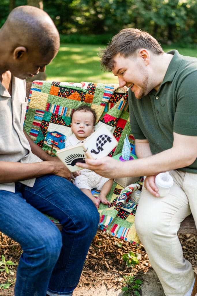 Two dads go through a book with their baby son while sitting on a bench.