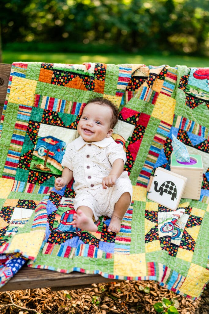 A baby boy sits on a handmade quilt draped on a bench and smiles.
