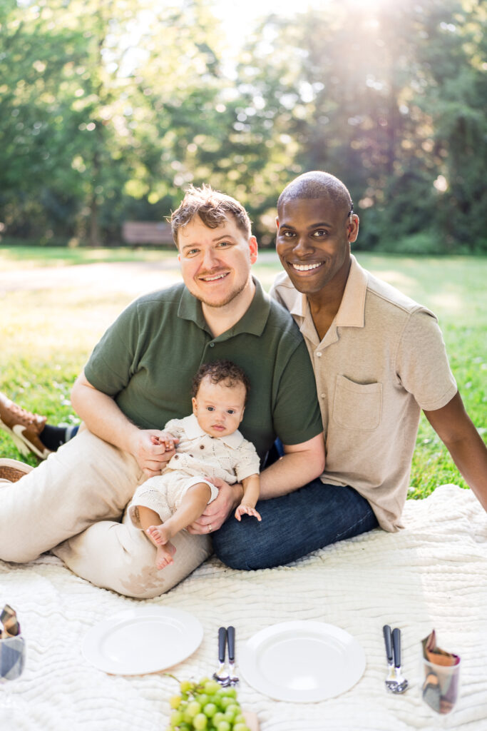 Two dads hold their baby son in an outdoor picnic setting.