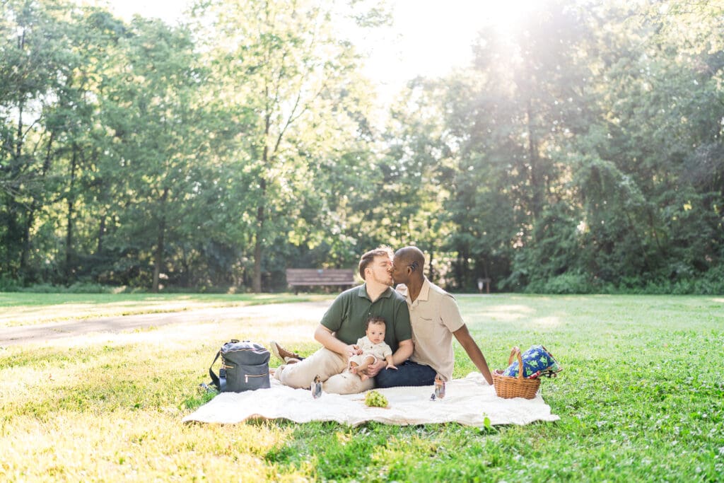 Two dads kiss while hold their baby son in an outdoor picnic setting.