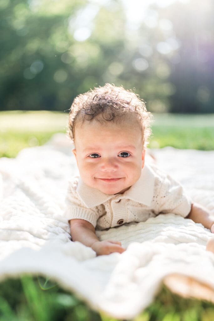 A baby boy smiling while laying on his stomach on a blanket outside.