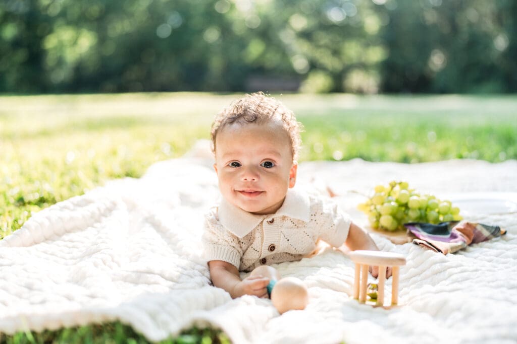 A baby boy smiling while laying on his stomach on a blanket outside.