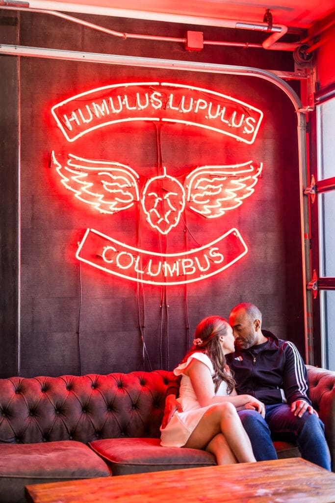 Bride and groom in casual outfits sit on a couch in front of a glowing sign at BrewDog in Columbus, Ohio