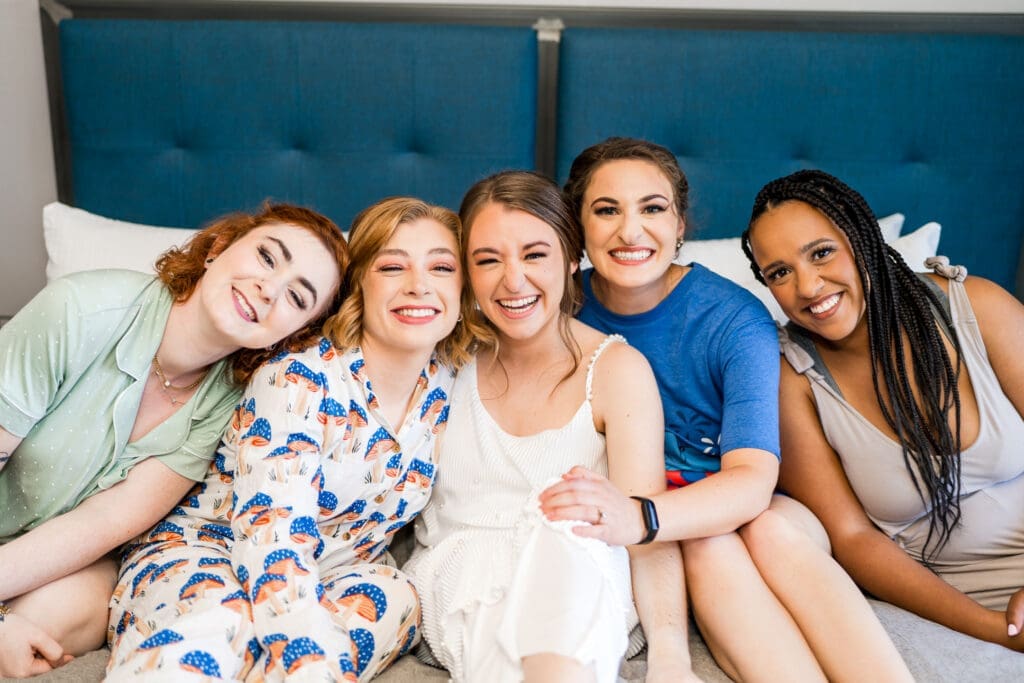 The bride and her bridesmaids in their getting ready outfits laughing and sitting on the bed