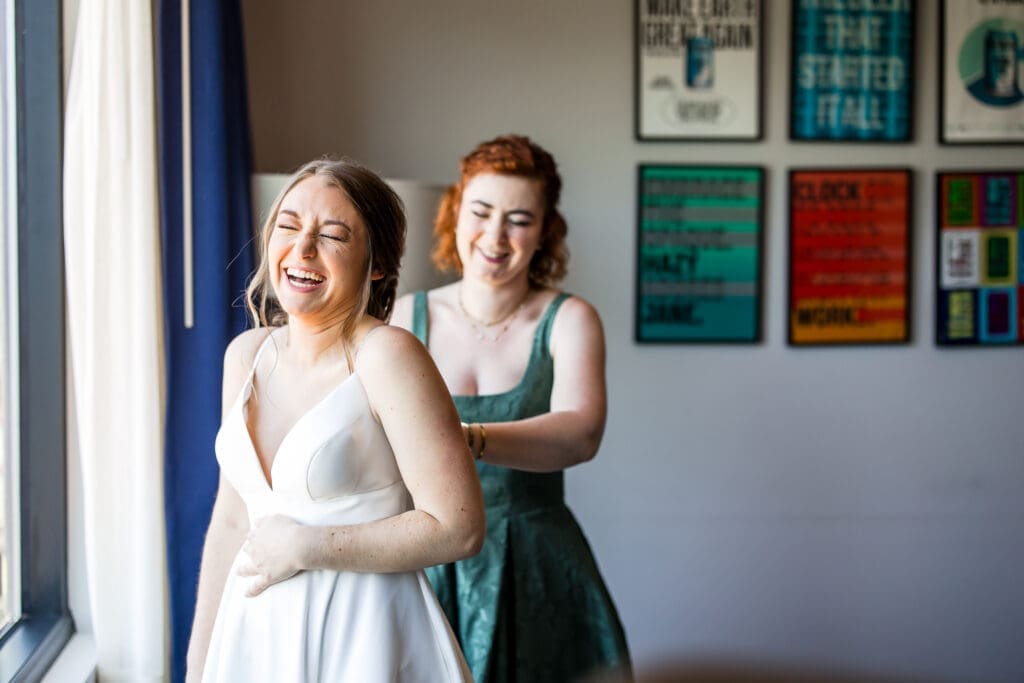 The bride laughs as her maid of honor buttons up her dress in Columbus, Ohio.